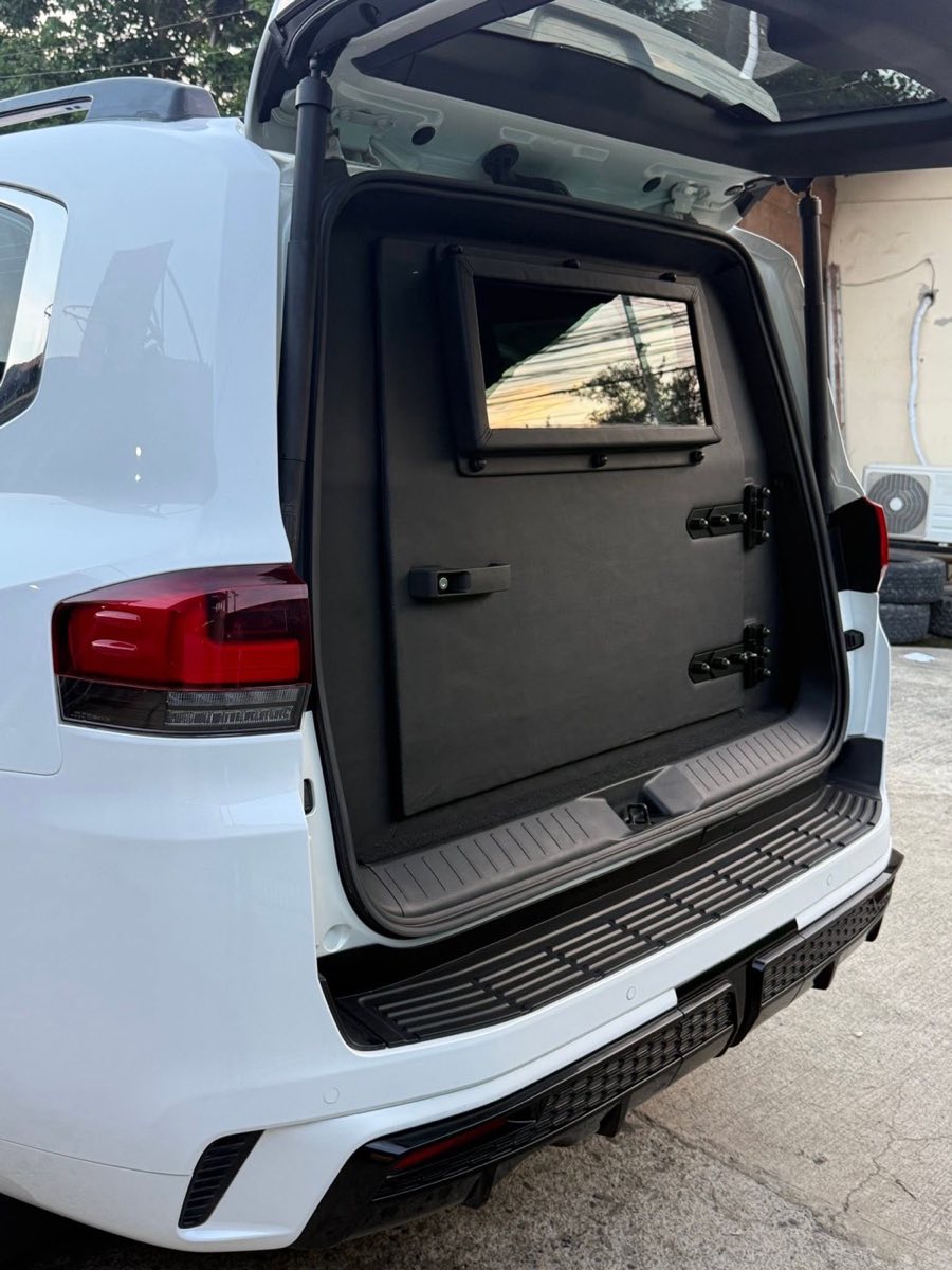 Armored rear door of a white Toyota Land Cruiser 300 open, showing the black ballistic panel and gun-port window